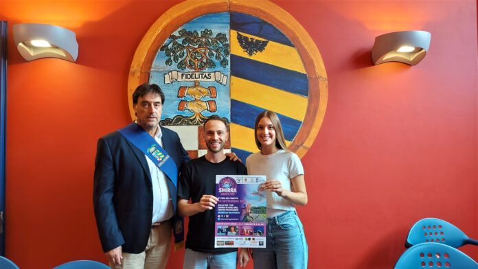 Tre persone posano davanti a uno stemma su una parete rossa, tengono un poster della manifestazione Smirra Games Fest.