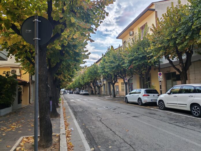 Strada urbana con alberi e auto parcheggiate lungo la via, in una giornata con cielo parzialmente nuvoloso.