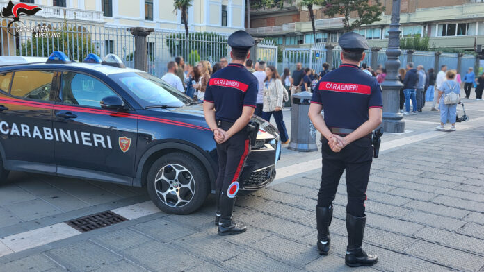 Carabinieri in divisa davanti all'ingresso di un edificio scolastico, con studenti e passanti sullo sfondo.
