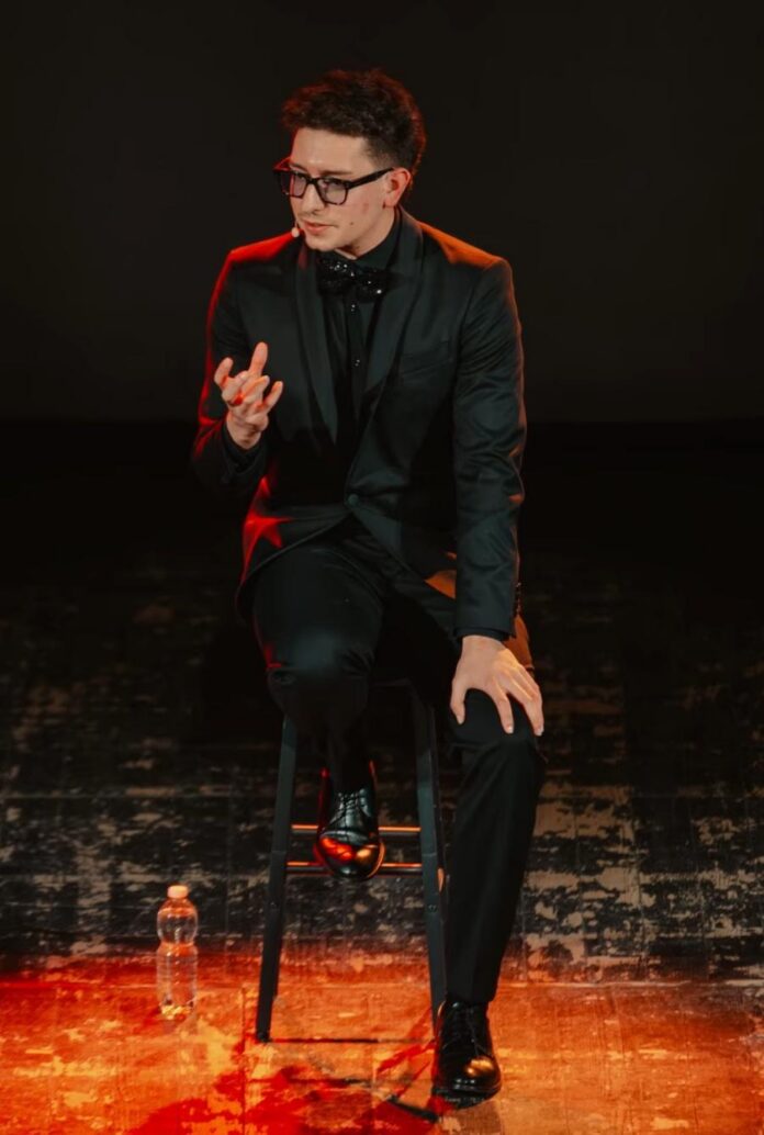 Il creator Shamzy seduto su uno sgabello sul palco con abito nero e cravatta verde, mentre gesticola durante uno spettacolo di stand-up comedy.