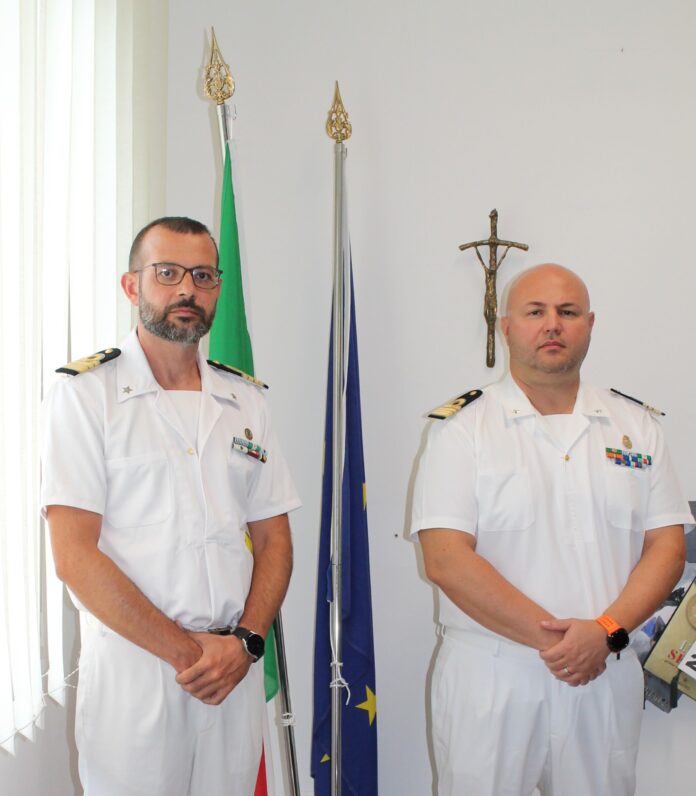 Due ufficiali della Capitaneria di Porto in uniforme bianca in piedi davanti a bandiere e un crocifisso.