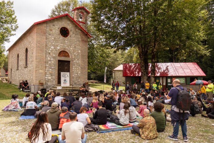 Gruppo di persone in un bosco della Sila Piccola, un uomo con giacca gialla e cappello bianco parla al microfono mentre i partecipanti ascoltano seduti o in piedi.