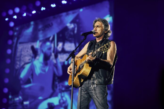 Luciano Ligabue in concerto con chitarra e microfono, sfondo di luci blu e proiezione grande.