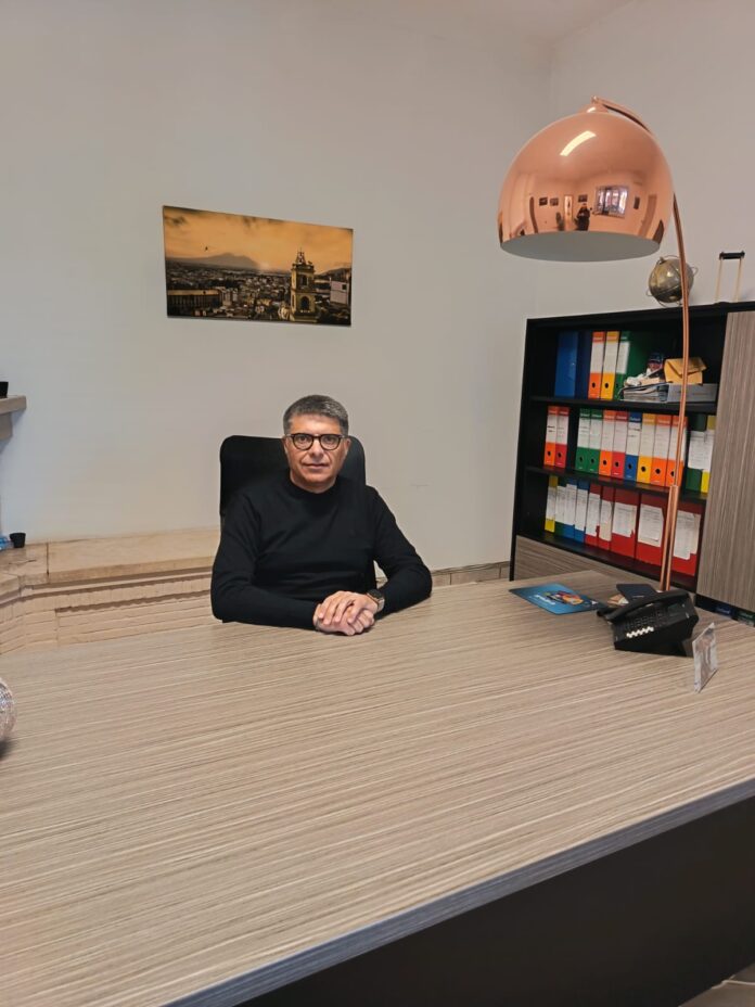 Aniello Buonaiuto seduto alla scrivania di un ufficio con mensole e lampada.