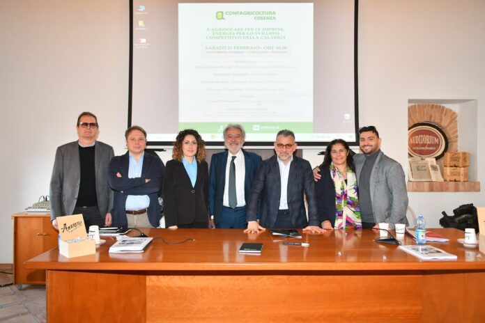 Relatore parla al microfono durante un convegno sull'energia, con altre persone sedute a un tavolo sullo sfondo.