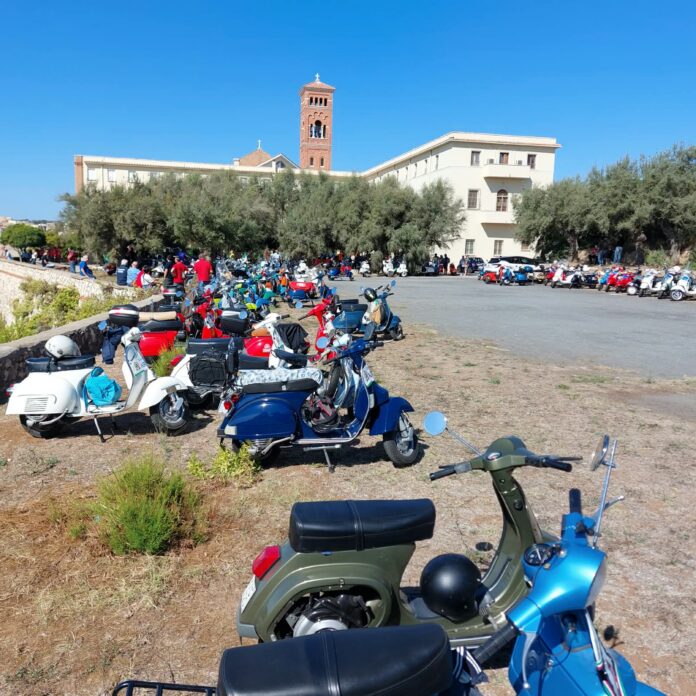 Raduno di Vespa con molti scooter parcheggiati all'aperto e persone riunite davanti a un edificio storico.