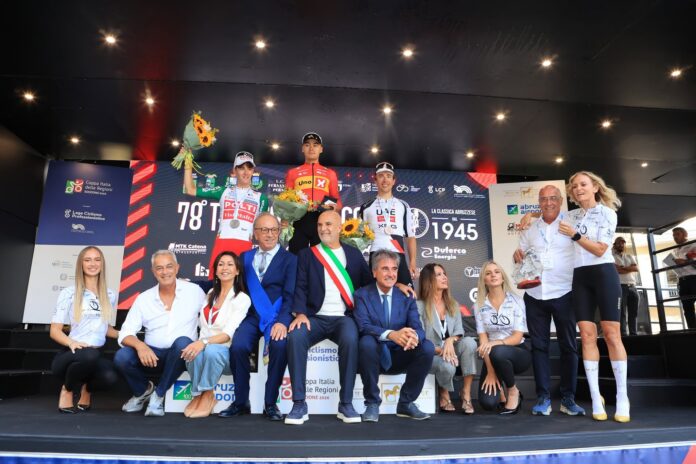 Gruppo di ciclisti in corsa durante il 78° Trofeo Matteotti su strada asfaltata con pubblico ai lati e segnali stradali.