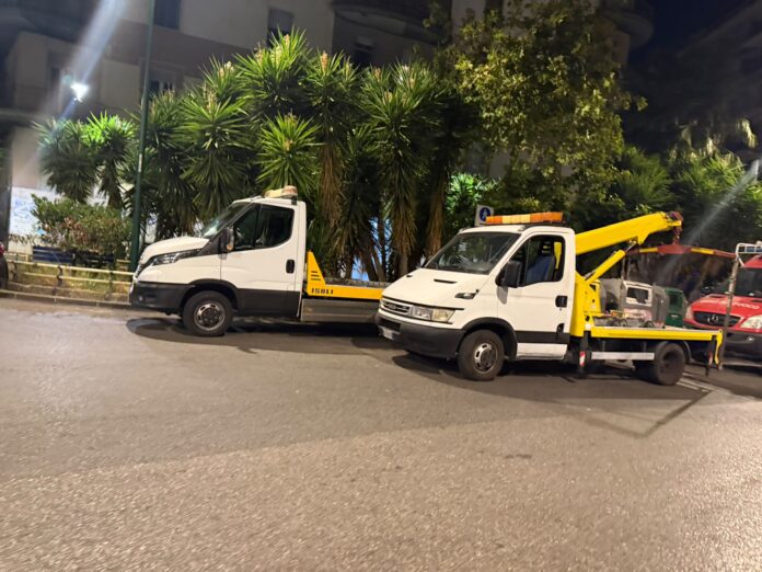 Due carri attrezzi bianchi parcheggiati di notte in una strada urbana con alberi e palazzi sullo sfondo.