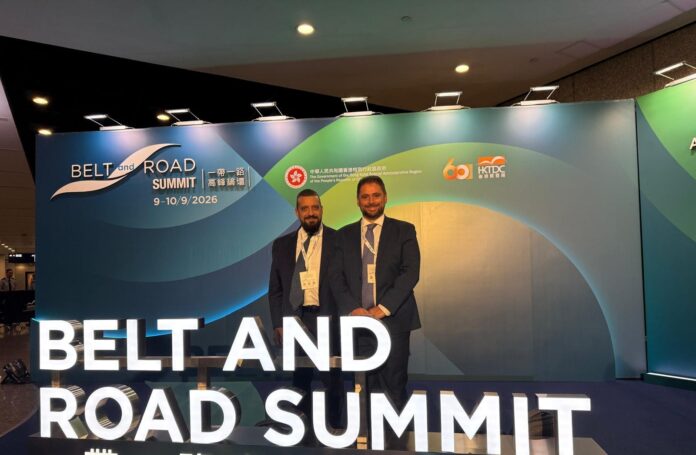 Due uomini in abito formale posano davanti allo stand del Belt and Road Summit 2026 a Hong Kong.