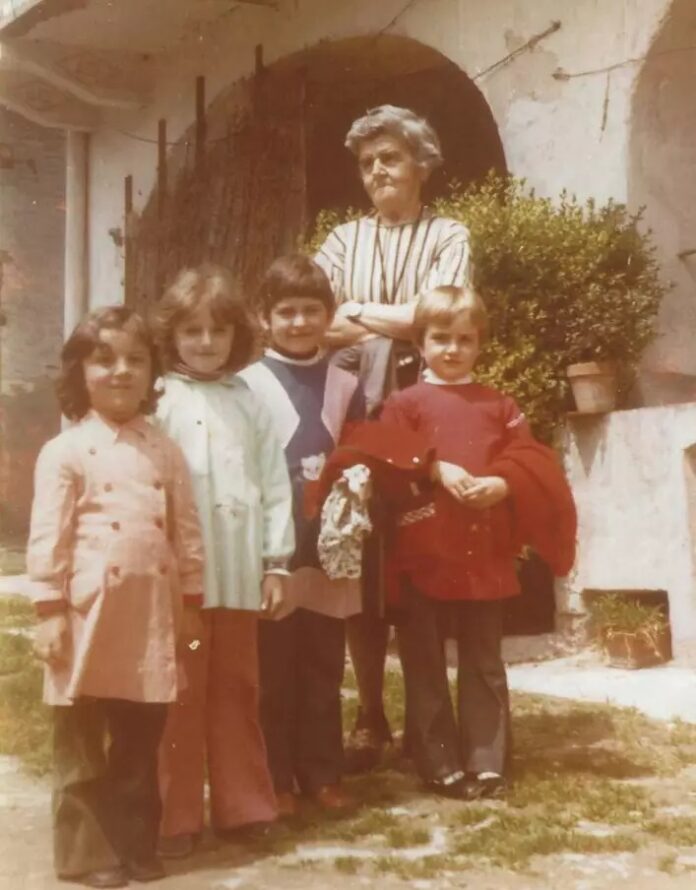 Foto storica di una maestra con quattro bambini all'aperto, in un cortile con un arco sullo sfondo.