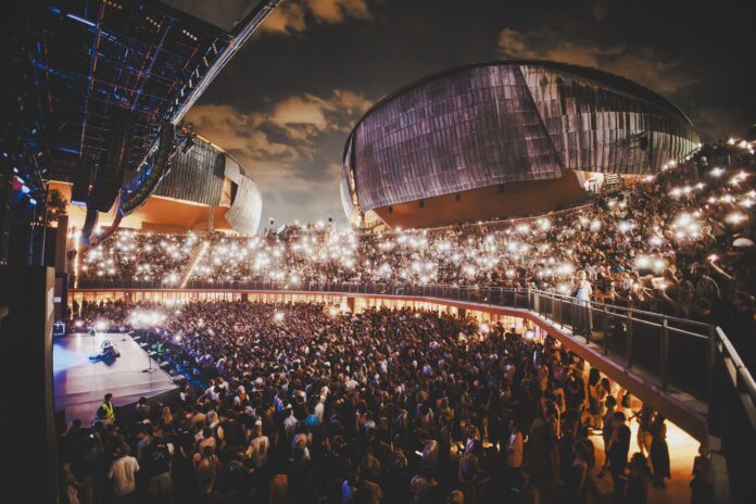 Veduta notturna dell'Auditorium Parco della Musica con moltissime persone sedute e in piedi durante un concerto.