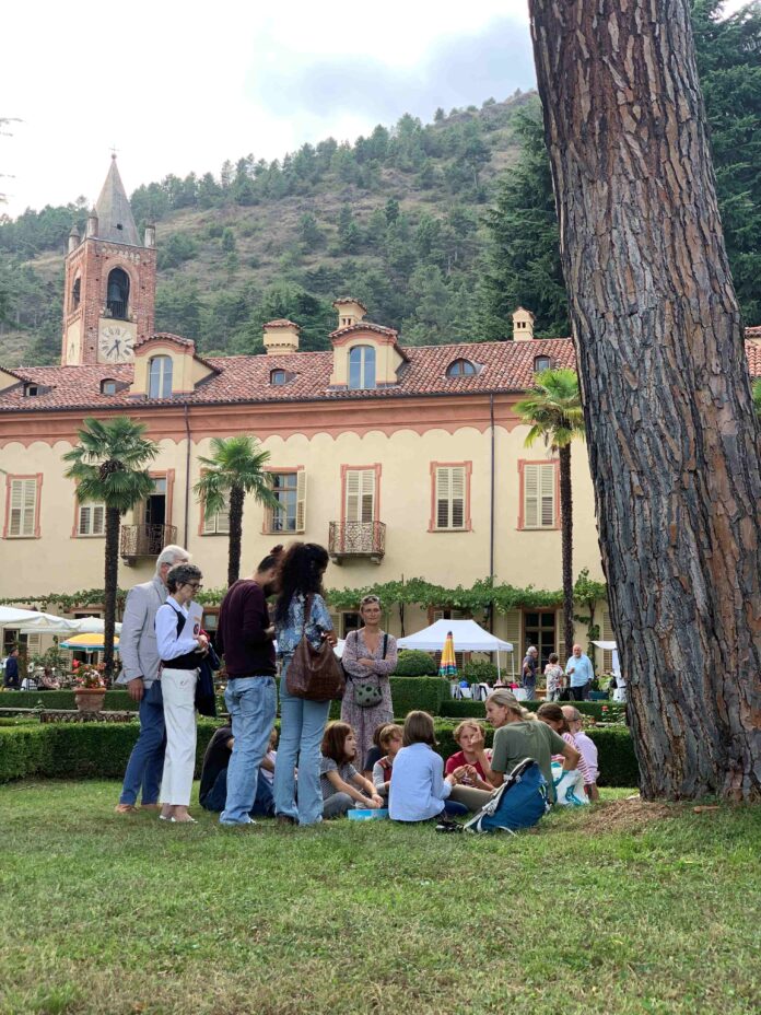 Persone sedute e in piedi che partecipano a un'attività nel giardino con piante e fiori, con muro in fondo.