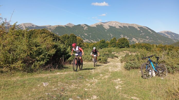 Due ciclisti in mountain bike pedalano su un sentiero sterrato con montagne sullo sfondo, immagine illustrativa per la Granfondo Parco Nazionale d’Abruzzo.