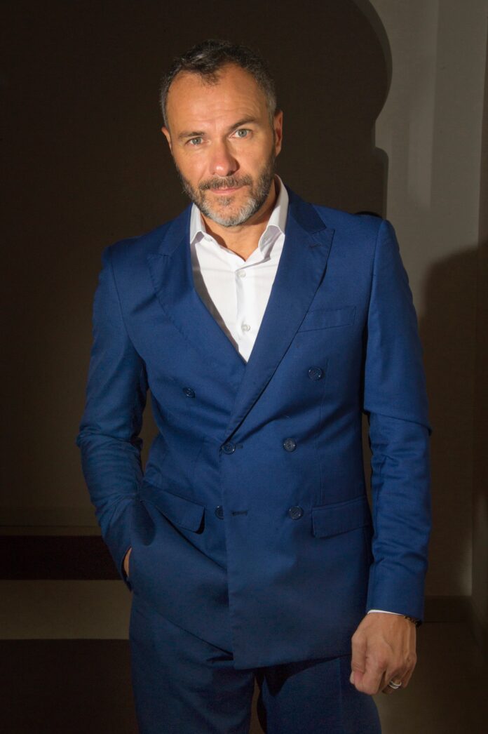 Massimiliano Gallo ritratto in abito blu in ambiente con illuminazione soffusa.