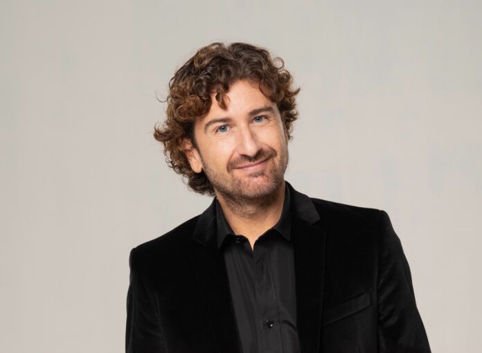 Ritratto di Alessandro Siani con giacca nera e camicia nera, su sfondo neutro.