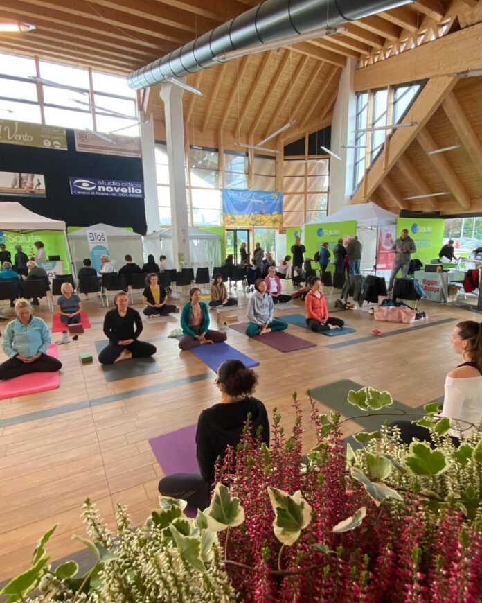 Persone sedute su tappetini per attività di benessere nel Palazzetto di Canove, Roana, durante evento wellness.