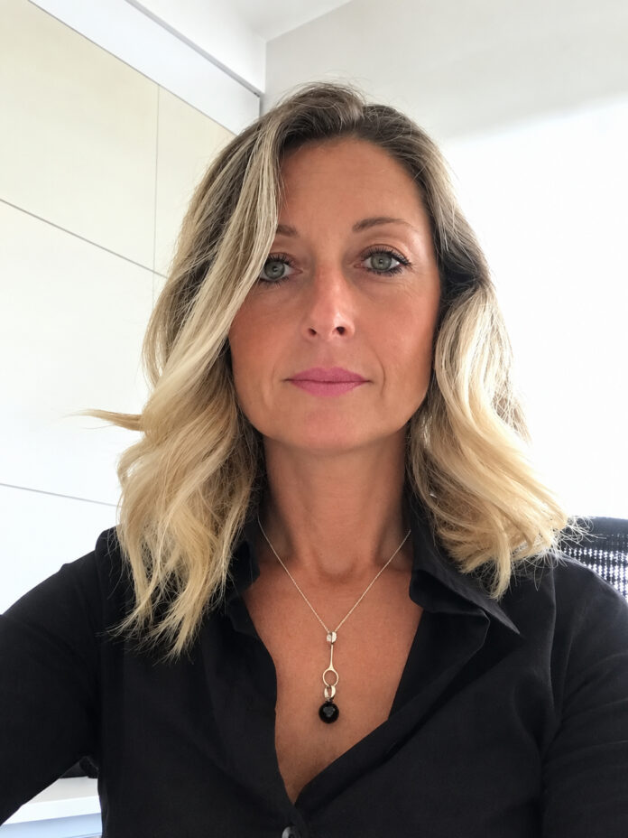 Ritratto fotografico di una donna sorridente con capelli biondi e ondulati, indossa una camicia nera e una collana.