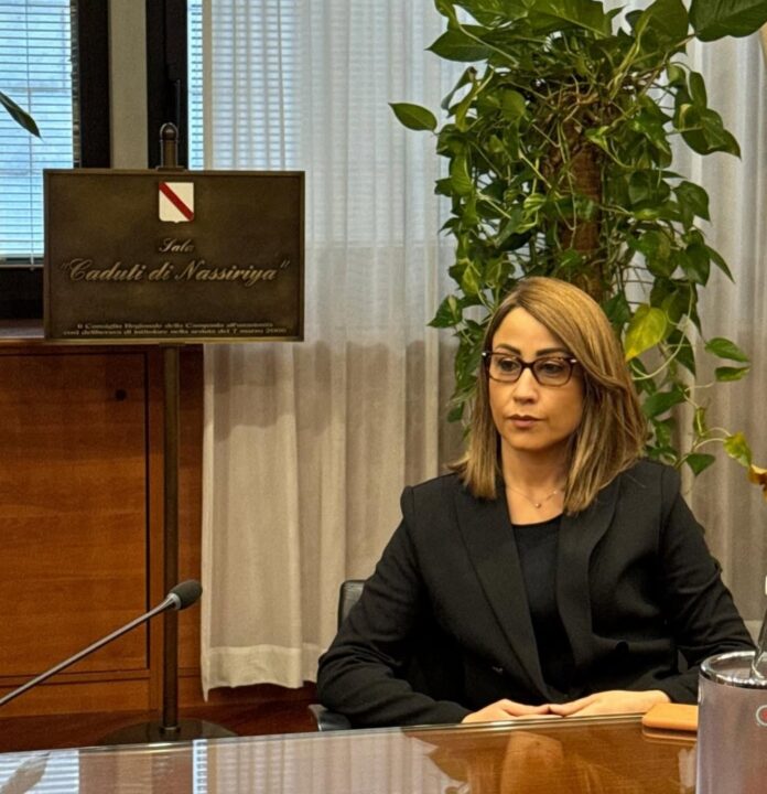 Consigliera Michela Rostan seduta a un tavolo in ambiente istituzionale con una targa commemorativa sullo sfondo.