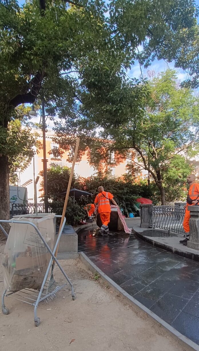 Operatori con divise arancioni al lavoro per il lavaggio e la rimozione di rifiuti in una piazza cittadina con panchine e alberi.