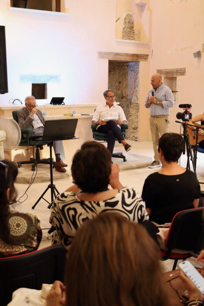 Interno di un palazzo con persone sedute che assistono a una conferenza o presentazione, con tre persone in primo piano, una delle quali parla al microfono.