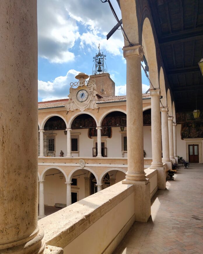 Orologio storico a torre nel cortile interno di un palazzo storico con archi e colonne, quadro con lancetta in cerchio e campane sulla torretta.