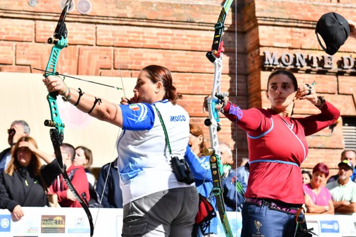 Arcieri che tirano con arco durante una gara all'aperto con arco e frecce, con arco gonfiabile della Federazione Italiana Tiro con l'Arco sullo sfondo.