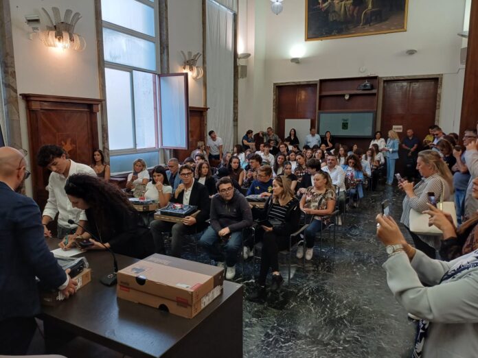 Cerimonia di consegna computer portatili agli studenti in una sala affollata con persone sedute e alcune persone che ritirano i PC su un tavolo.