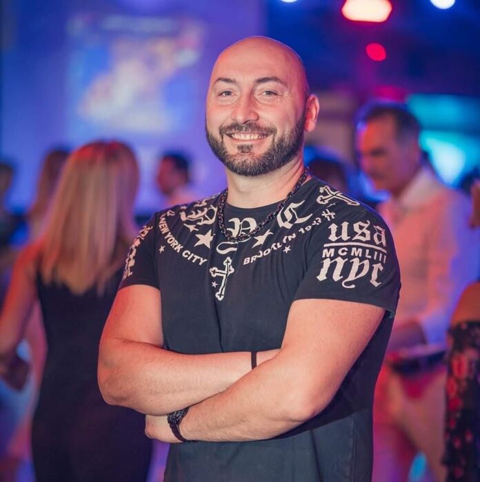 Paolo Citro sorridente con maglietta nera, in un ambiente con luci colorate e persone sullo sfondo, in una foto illustrativa legata a 