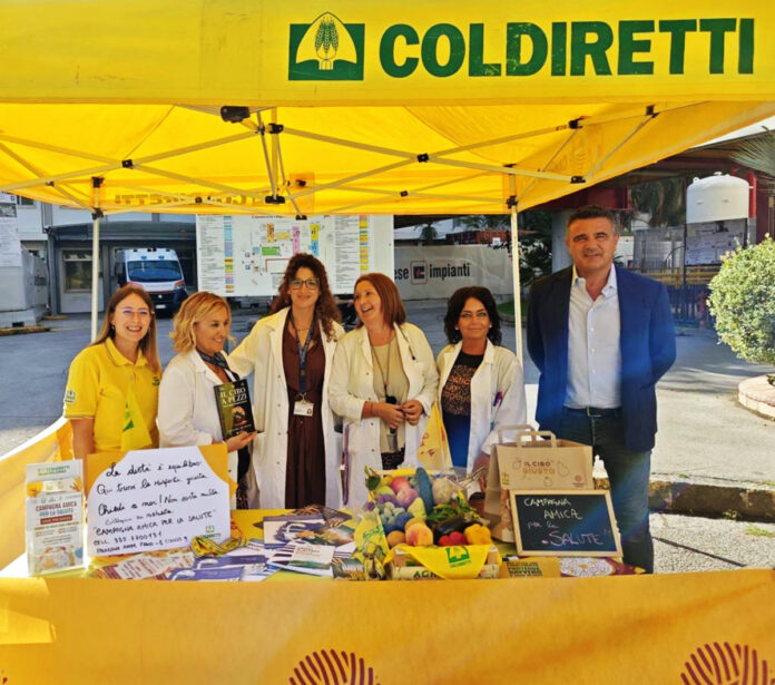 Stand di Campagna Amica con persone e materiale informativo sotto gazebo giallo in un contesto esterno luminoso.