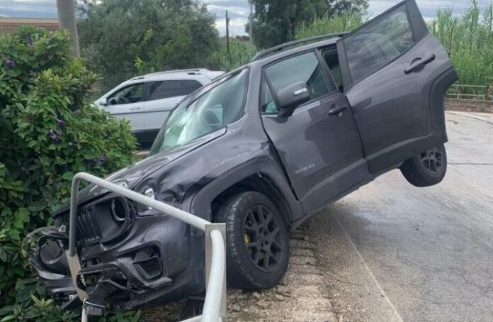 Auto grigia sospesa sul bordo di una strada con danni e barriere di protezione piegate, immagine illustrativa incidente stradale.
