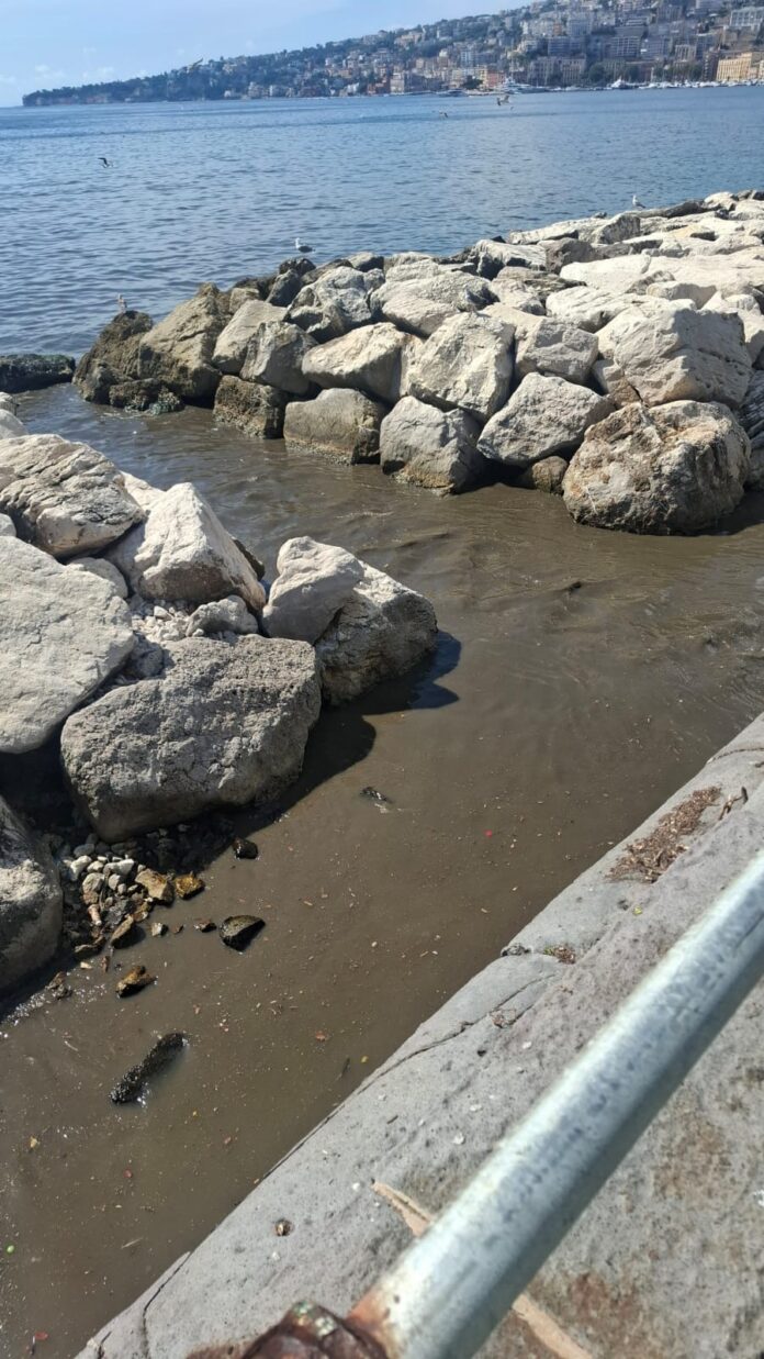 Sversamento di acqua putrida maleodorante tra le rocce sul litorale di Via Caracciolo, Napoli, con mare visibile sullo sfondo.