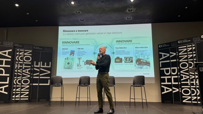 Persona che parla in un auditorium davanti a una presentazione proiettata con il logo Alpha Innovation e contenuti relativi all'innovazione di Fujifilm.