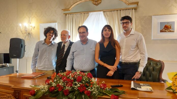Cerimonia di premiazione con cinque persone dietro un tavolo con composizione floreale in primo piano, in una sala con pareti decorate.