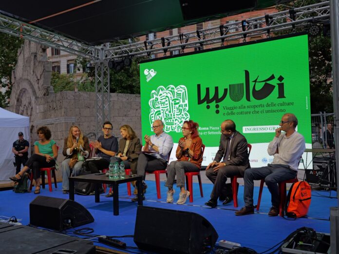 Panel di relatori seduti su palco con sfondo schermo verde con scritte del Festival Multi a Piazza Vittorio.