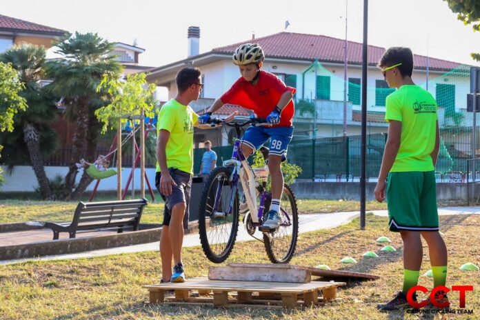 Un ragazzo in tenuta da ciclismo mentre salta con la bicicletta su un ostacolo, assistito da due persone in magliette gialle all'aperto in un parco.
