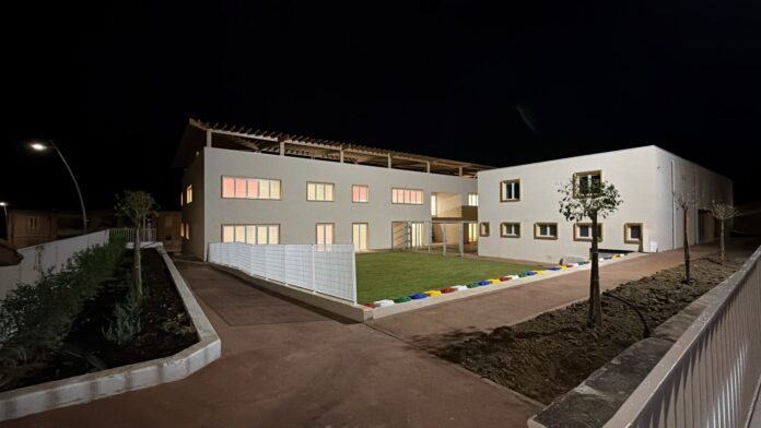 Edificio scolastico illuminato di sera con area verde e vialetto d'ingresso.