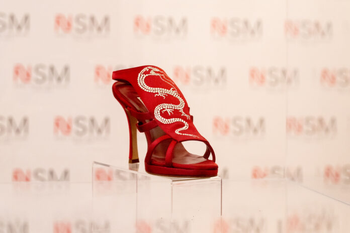 Scarpa rossa decorata con strass esposta su un piedistallo trasparente su sfondo sfocato con logo NSM.