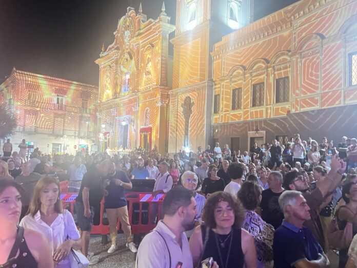 Piazza di Frattamaggiore illuminata con videomapping e folla durante la festa patronale con musica popolare.