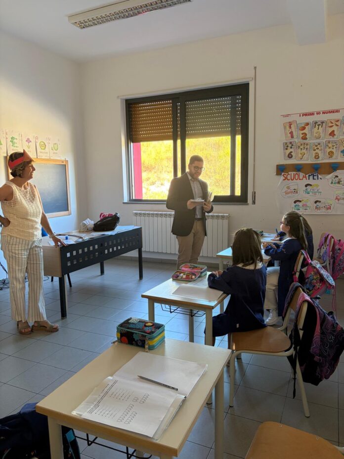 Classe scolastica con studenti seduti ai banchi e insegnanti o amministratori in piedi in aula durante il primo giorno di scuola.