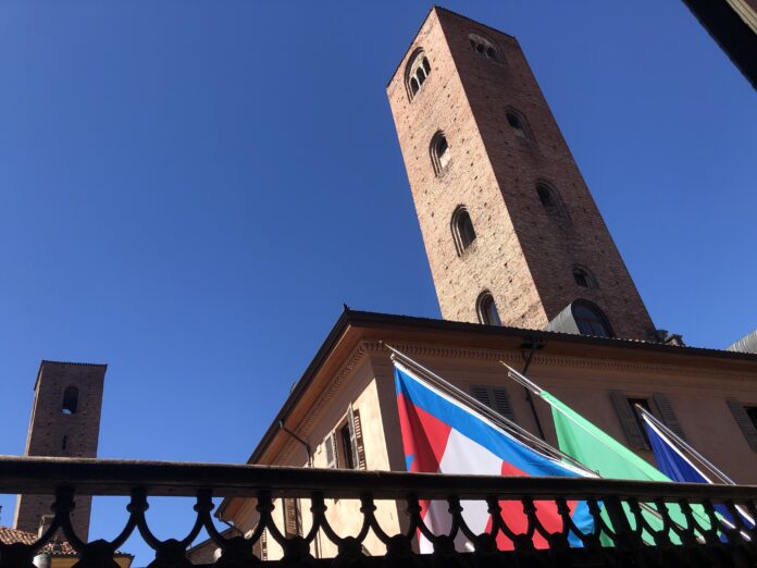 Veduta di una torre storica ad Alba con alcune bandiere visibili in primo piano.