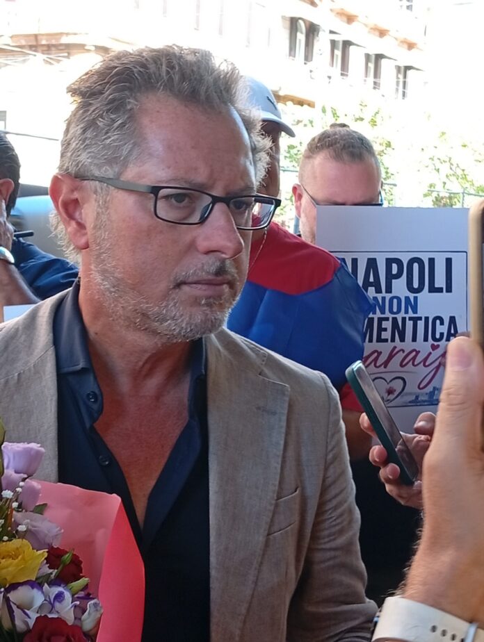 Persone durante una manifestazione con fiori e un simbolo commemorativo in zona urbana di Napoli, in ricordo di una giovane vittima.