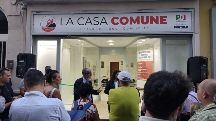 Persone radunate davanti all'ingresso di La Casa Comune con il logo visibile sopra la porta durante l'inaugurazione a Bacoli.