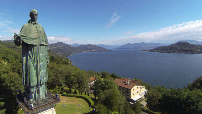 Statua di San Carlo Borromeo ad Arona con panorama del Lago Maggiore sullo sfondo, prato sottostante e cielo sereno.