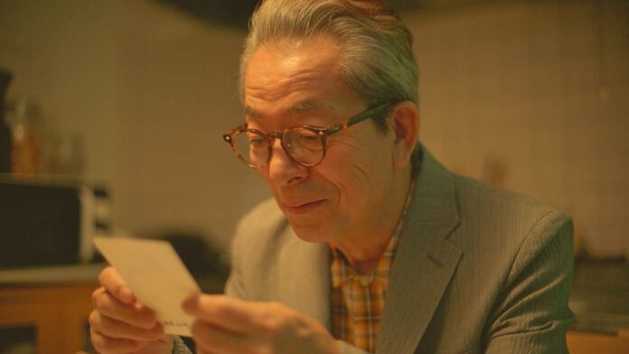 Primo piano di Yutaka Mizutani con occhiali e maglietta scura su sfondo blu.