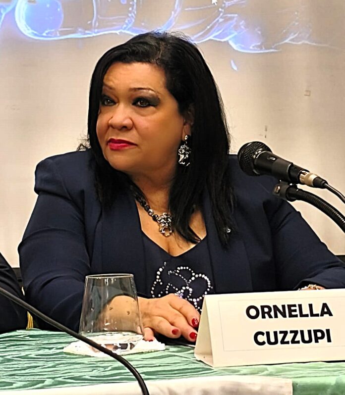 Donna seduta a un tavolo con microfono e cartellino con nome Ornella Cuzzupi, durante un evento o conferenza.
