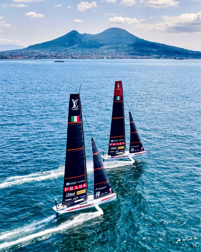 Due barche da regata con vele nere nel Golfo di Napoli, con il Vesuvio sullo sfondo.