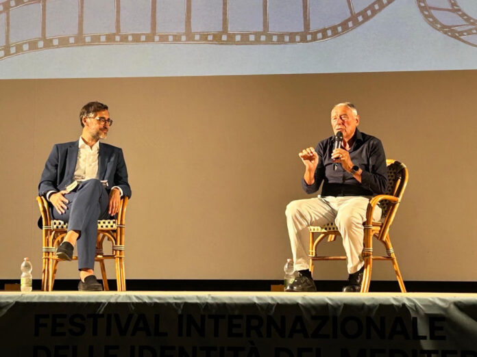 Incontro su palco con due uomini seduti su sedie di fronte a un grande schermo con scritta Pietro Grasso 'U Maxi durante il Festival Internazionale delle Identità del Mediterraneo a Marzamemi.