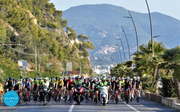 Partecipanti della Granfondo Internazionale Alassio in gruppo al via sulla strada con mare e palme sul lato.