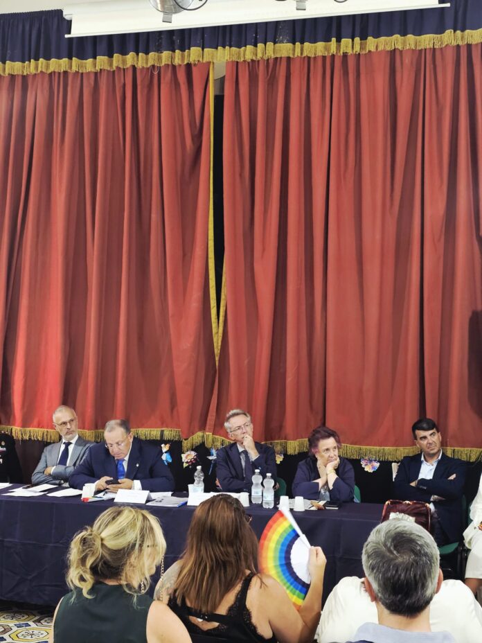 Incontro pubblico in una sala con persone sedute di fronte a un palco con tavolo e microfoni, decorazioni colorate e un grande tendone rosso sullo sfondo.