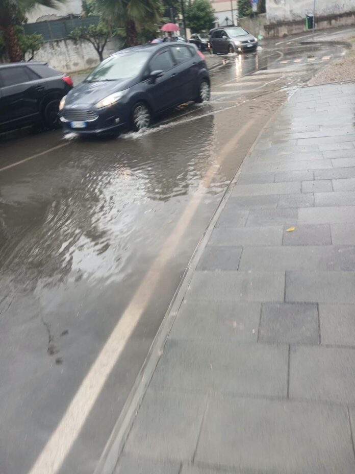 Strada allagata con auto che attraversano pozzanghere a San Marzano sul Sarno, immagine illustrativa del disagio per tombini ostruiti.